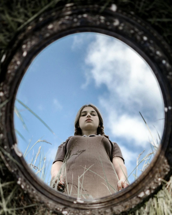 a woman standing in a field looking at herself in a mirror, by Lucia Peka, looking at us from a porthole, maisie williams, shot from below, trending photo