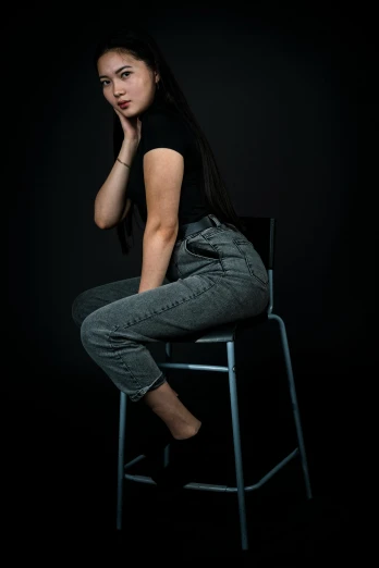 a woman sitting on a stool in the dark, a character portrait, by irakli nadar, unsplash, wearing jeans, girl with black hair, studio medium format photograph, candid!! dark background