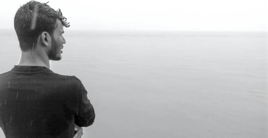 a man standing on top of a beach next to the ocean, a black and white photo, romanticism, header, frank dillane, fog!, panoramic view of girl