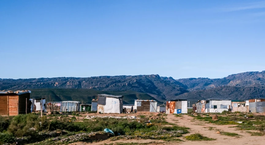 a dirt road with shacks and mountains in the background, by Hubert van Ravesteyn, unsplash, renaissance, makeshift houses, coast, standing in township street, panoramic