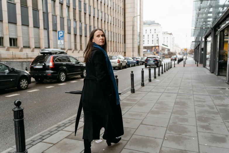 a woman standing on a sidewalk in front of a building, by Emma Andijewska, wearing a long coat, photographed for reuters, standing bravely on the road, kirsi salonen