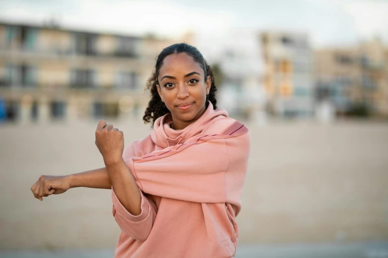 a woman is posing for a picture on the beach, an album cover, by Lily Delissa Joseph, pexels contest winner, happening, large fists, wearing a pink hoodie, alexandria ocasio-cortez, bandage on arms