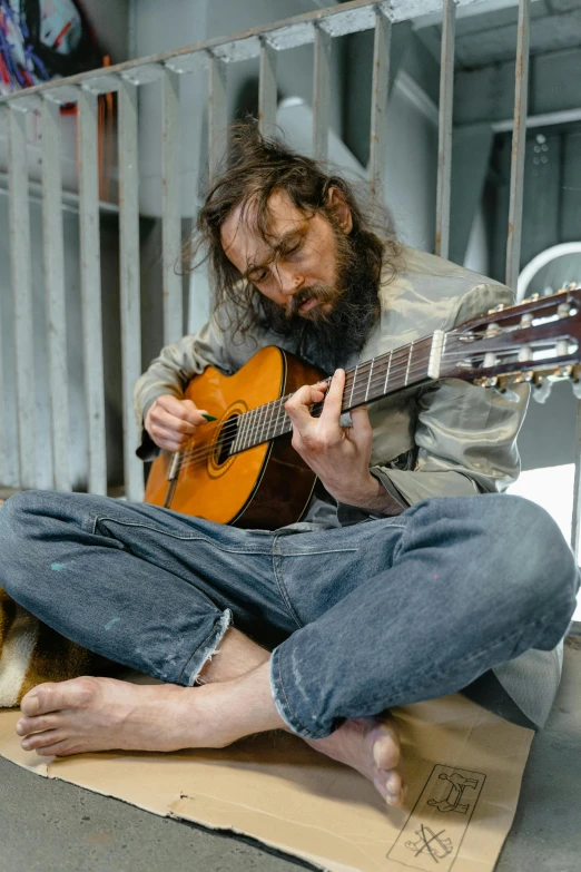 a man sitting on the ground playing a guitar, stood in a cell, bearded, scruffy looking, homeless