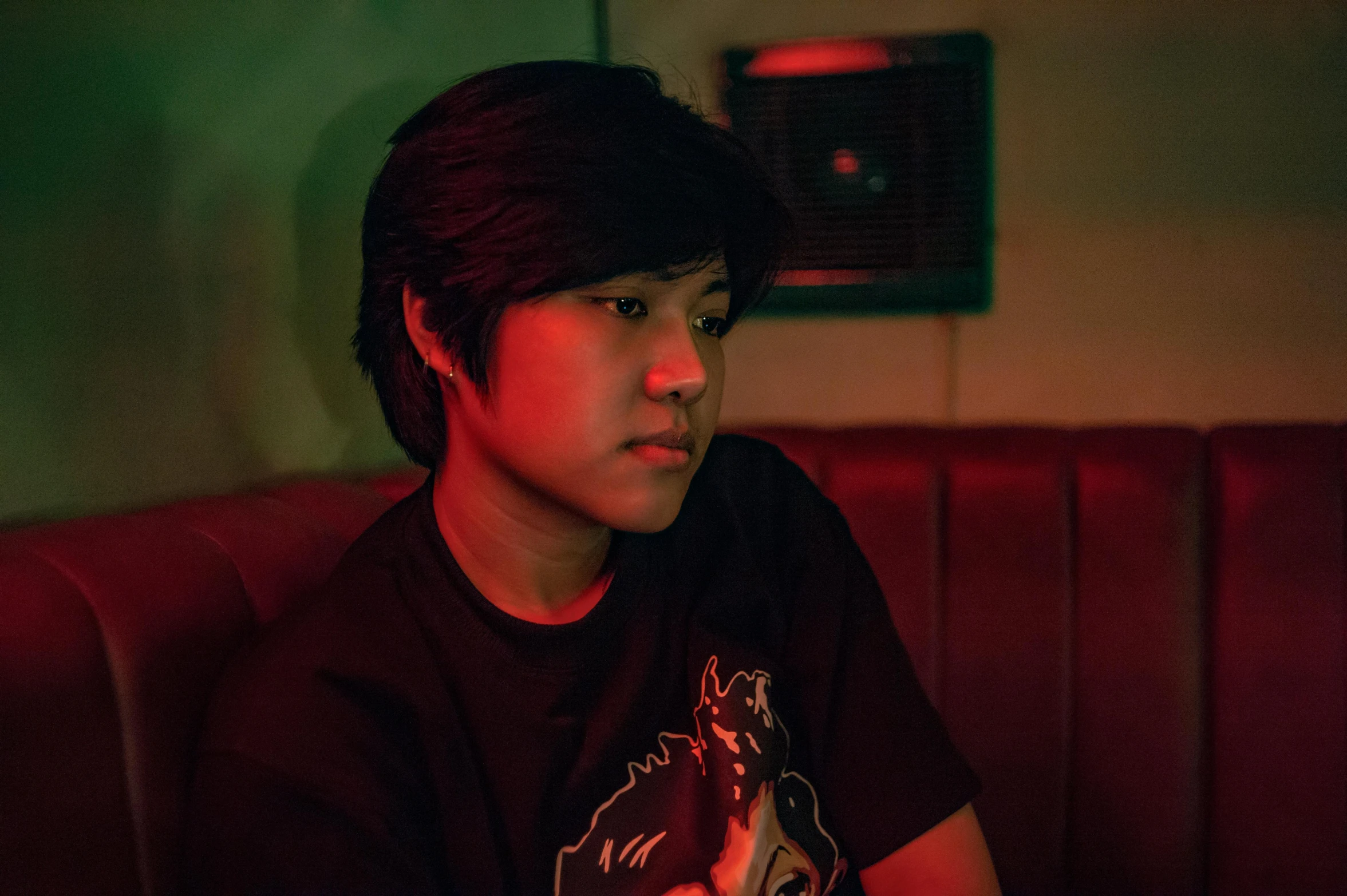 a boy sitting on a couch with a remote in his hand, inspired by Liam Wong, unsplash, realism, 8 0 s asian neon movie still, headshot profile picture, standing in a dimly lit room, he is wearing a black t-shirt