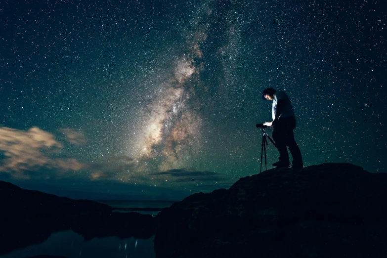 a man standing on top of a rock next to a body of water, pexels contest winner, many stars in the night sky, star walk, a photo of a man, like a catalog photograph