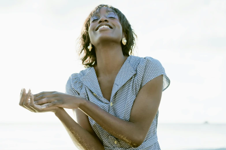 a woman standing on top of a sandy beach, earing a shirt laughing, naomi campbell, perspective from below, relaxed. blue background