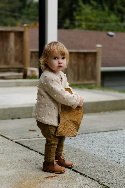 a little girl standing on a sidewalk holding a basket, by Winona Nelson, pexels contest winner, happening, brown jacket, gilded outfit, pouty, concrete