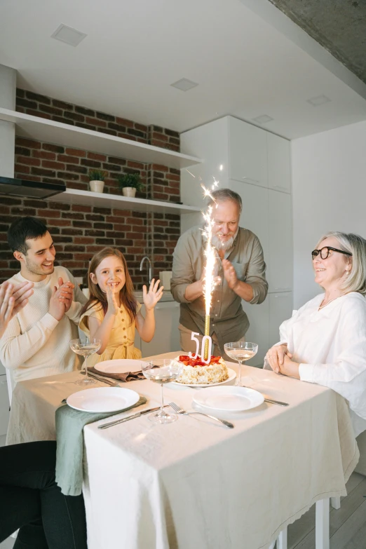 a group of people sitting around a table with a cake on it, white candles, happy family, sparkling light, aging