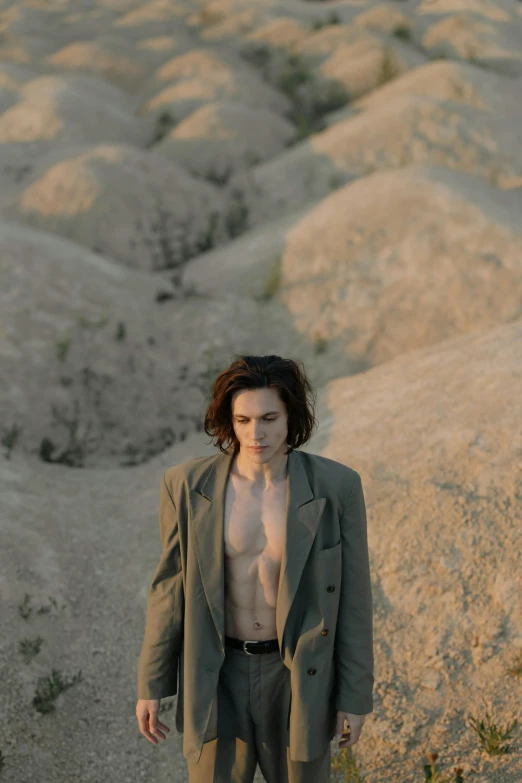 a man standing in the middle of a desert, an album cover, androgynous vampire, wearing a worn out brown suit, adam ondra, jacket over bare torso