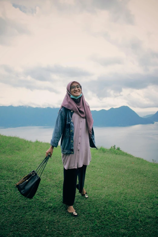 a woman standing on top of a lush green hillside, a picture, inspired by JoWOnder, sumatraism, hijab, fjords in background, wearing dirty travelling clothes, ((portrait))