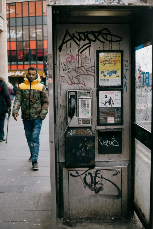 a phone booth sitting on the side of a street, a photo, by Sebastian Spreng, trending on pexels, graffiti, man standing, wearing a turtleneck and jacket, camo, jamel shabazz