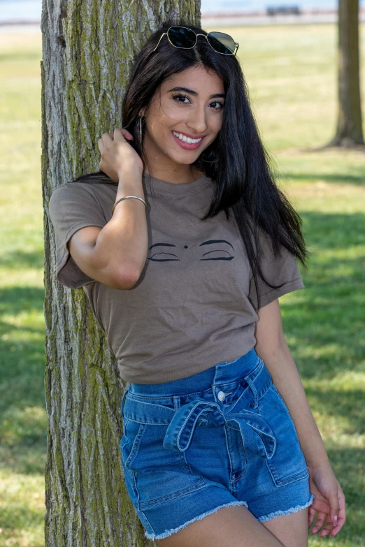 a woman leaning against a tree in a park, tan skin a tee shirt and shorts, smiling cat, mia khalifa, official store photo