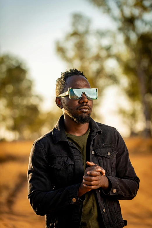 a man standing in a field wearing a pair of goggles, inspired by Albert Namatjira, trending on unsplash, afrofuturism, photo from a promo shoot, portrait photo of a backdrop, lit from the side, casually dressed