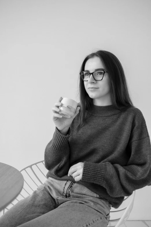 a woman sitting on a chair holding a coffee cup, a black and white photo, girl with glasses, andrea savchenko, wearing casual sweater, profile image