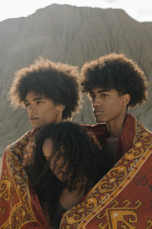 three people wrapped in a blanket in front of a mountain, an album cover, trending on pexels, afrofuturism, beautiful male twins portrait, big hair, black teenage boy, perfectly lit. movie still