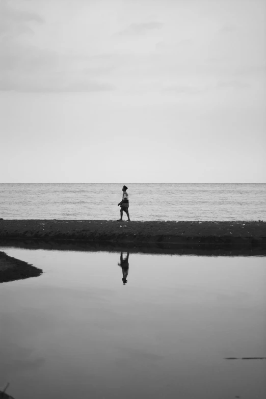 a person walking on a beach next to a body of water, a black and white photo, thawan duchanee, reflective water, run, road to the sea
