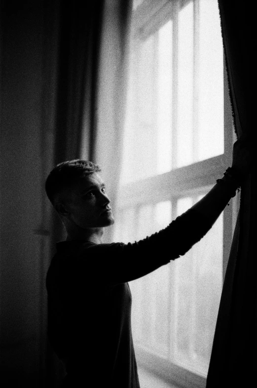 a man that is standing in front of a window, a black and white photo, unsplash, realism, curtain, yung lean, sunrise light, zayn malik