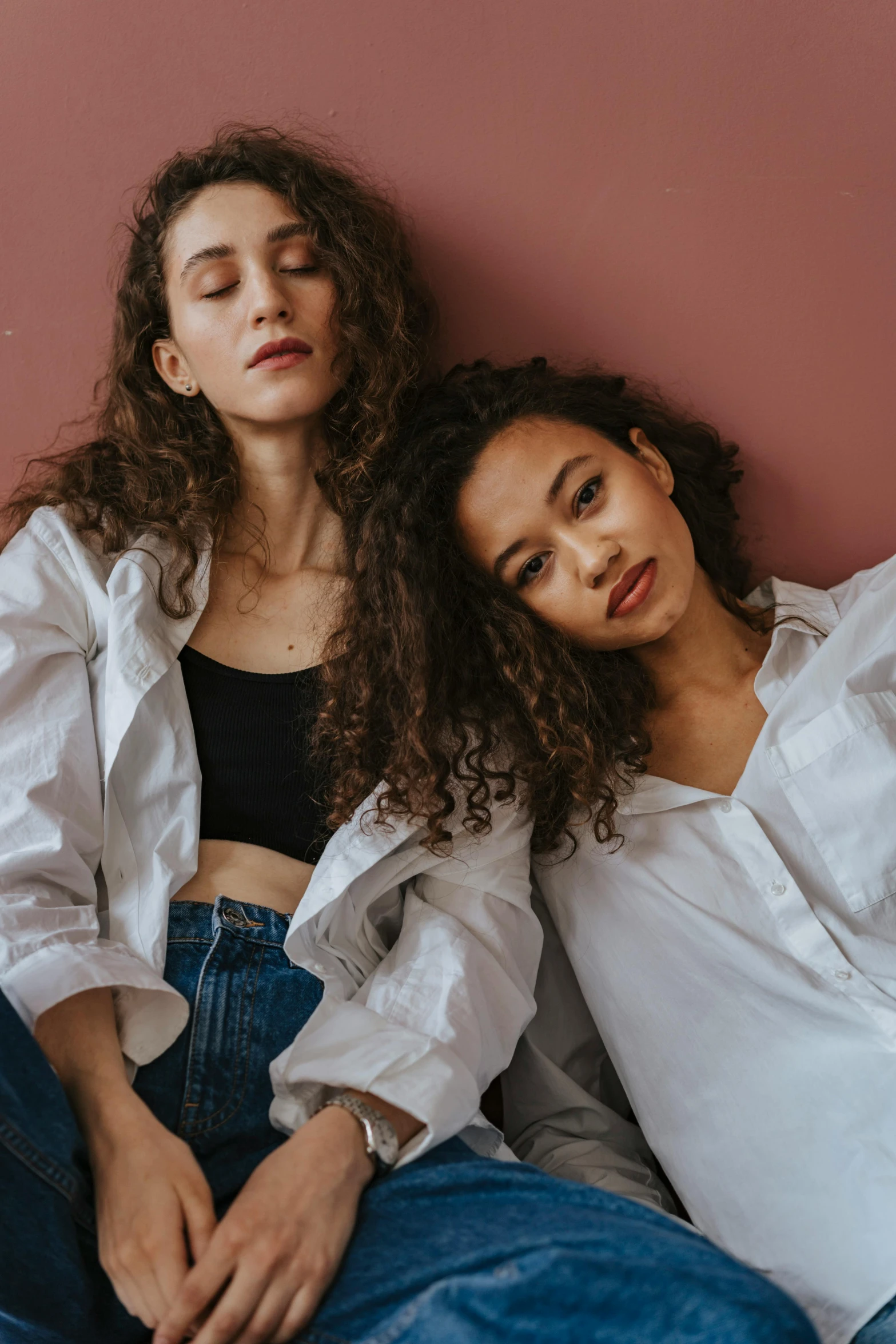 two women sitting next to each other on a bed, an album cover, trending on pexels, antipodeans, curls hair, wearing a white button up shirt, at a fashion shoot, bella poarch