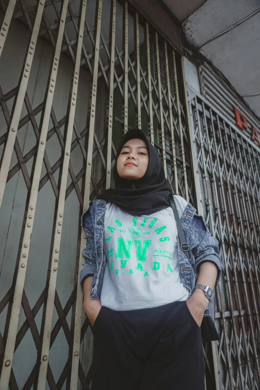 a woman standing in front of a metal fence, a picture, unsplash, hurufiyya, wearing a t-shirt, black and green, at college, studio picture