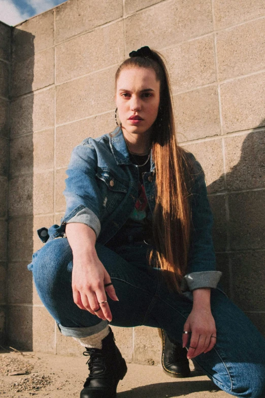a woman squatting in front of a brick wall, an album cover, by Gwen Barnard, unsplash, wearing a jeans jackets, portrait sophie mudd, long pigtail, androgynous male