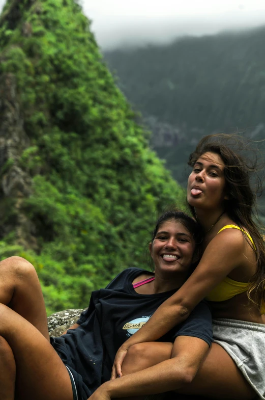 a couple of women sitting on top of a rock, in the background is lush jungle, tongue out, in between a gorge, beto val