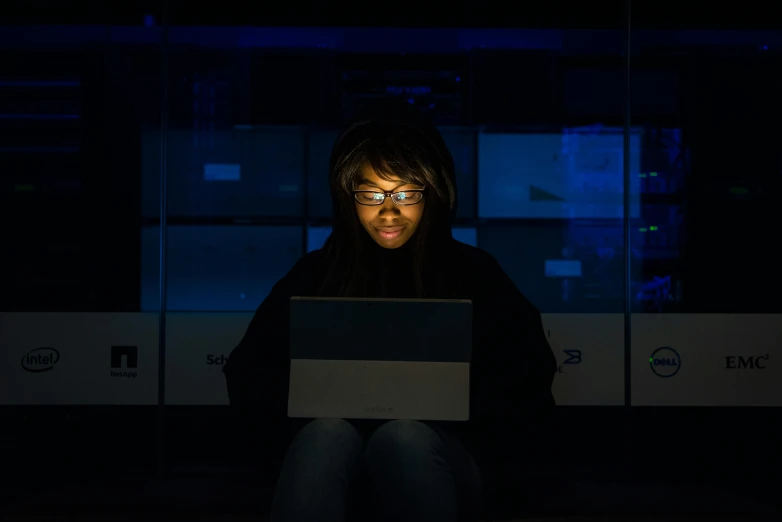 a woman sitting in the dark using a laptop, by Dennis Flanders, pexels, african female android, cold blue light, programming, avatar image