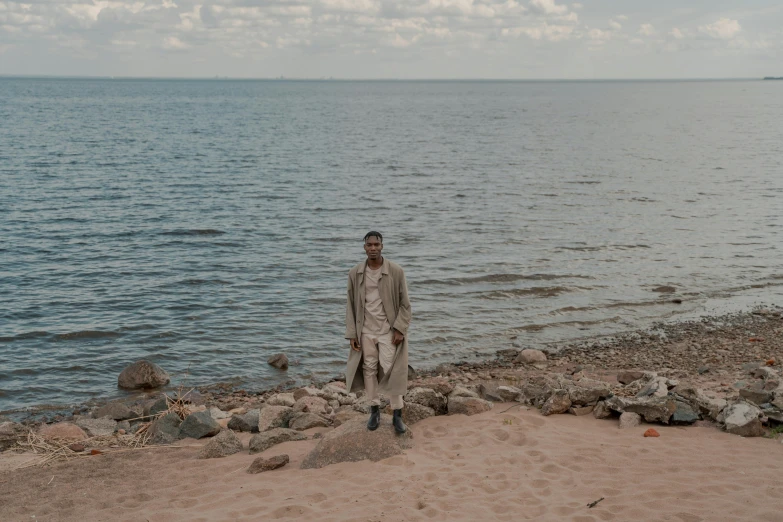 a man standing on a beach next to a body of water, an album cover, inspired by Oluf Høst, pexels contest winner, realism, wearing a long beige trench coat, helsinki, brown skinned, full-body-shot