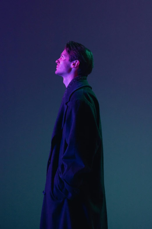 a man standing in front of a purple and blue background, an album cover, inspired by Russell Dongjun Lu, realism, he is wearing a black trenchcoat, grim fashion model looking up, standing in a dimly lit room, sideview