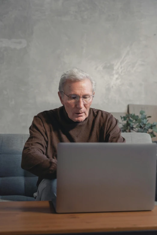 a man sitting in front of a laptop computer, by William Berra, trending on pexels, an oldman, on a gray background, he is wearing a brown sweater, people watching