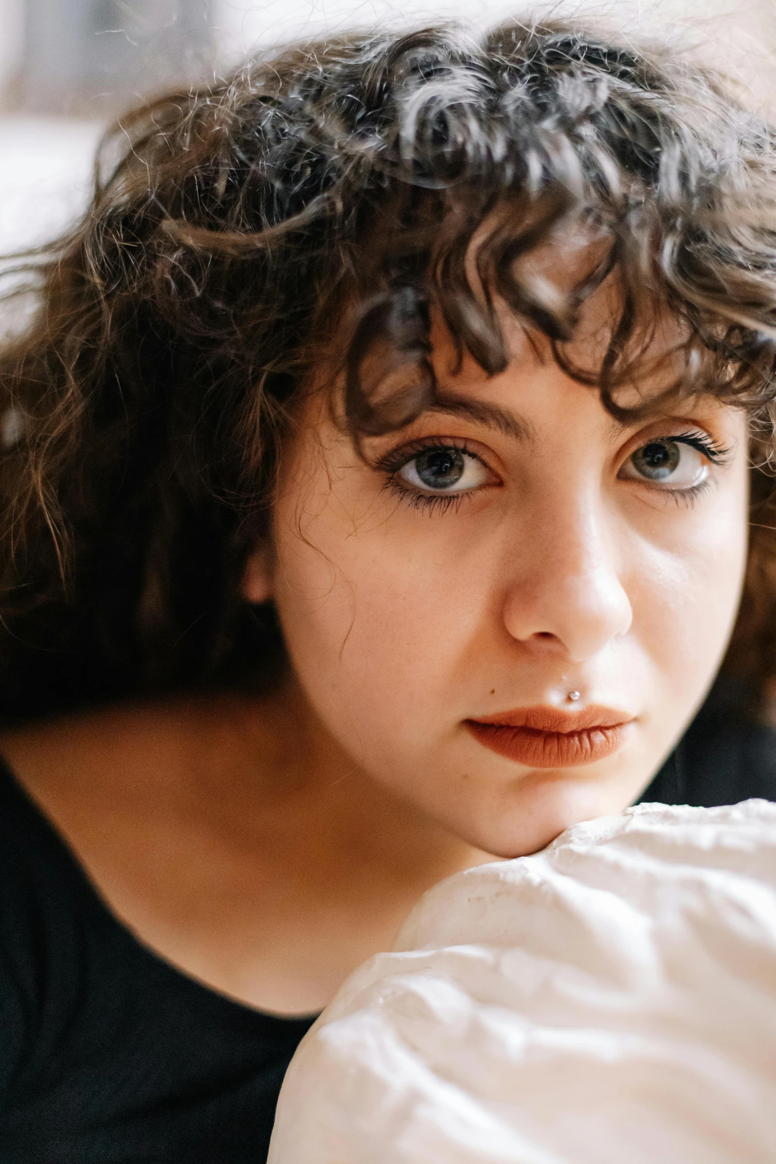 a woman sitting on top of a bed next to a pillow, an album cover, trending on pexels, renaissance, brown hair and large eyes, rebecca sugar, close up face female portrait, curly haired