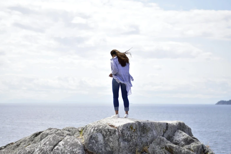 a woman standing on top of a rock next to the ocean, happening, wearing a linen shirt, high winds, blue, full shot photograph