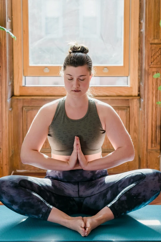 a woman sitting on a yoga mat in front of a window, by Jessie Algie, pexels contest winner, sitting with wrists together, promo image, liam brazier, wearing labradorite body armor