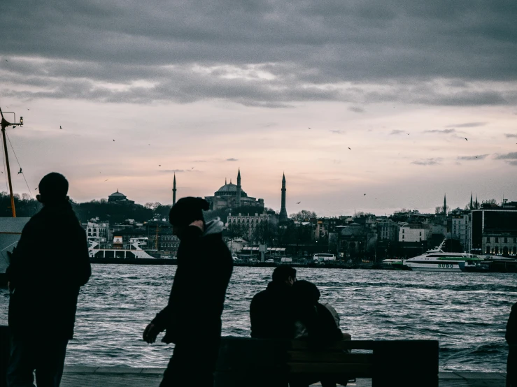 a group of people standing next to a body of water, by Ismail Acar, pexels contest winner, hurufiyya, istanbul, foreboding background, building in the distance, youtube thumbnail