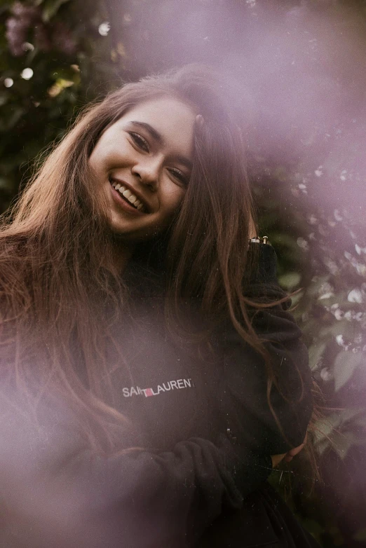 a woman smiling through the branches of a tree, by Sara Saftleven, unsplash, soft devil queen madison beer, wearing sweatshirt, ((purple)), teenager girl