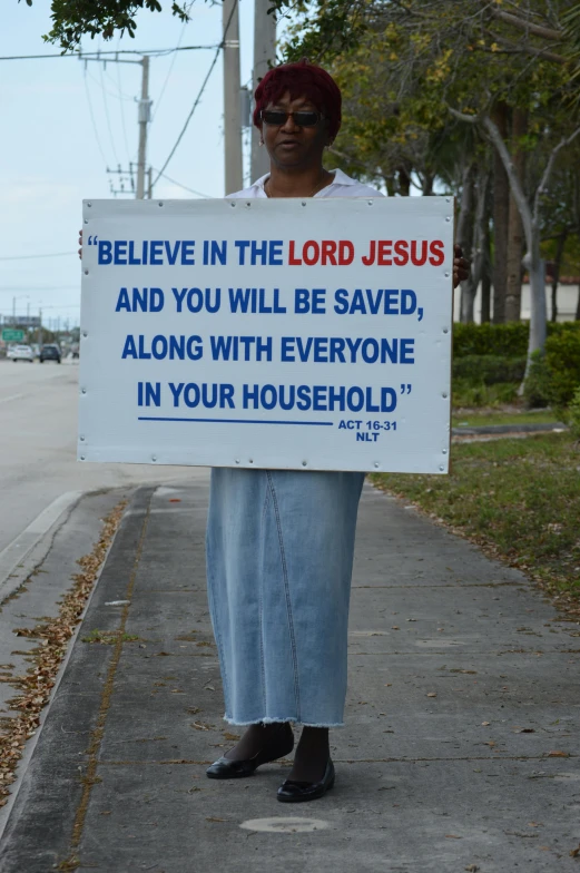 a woman holding a sign that says believe in the lord jesus and you will be saved along with everyone in your household, by Everett Warner, featured on reddit, miami beach, picture of a male cleric, b - roll, diverse