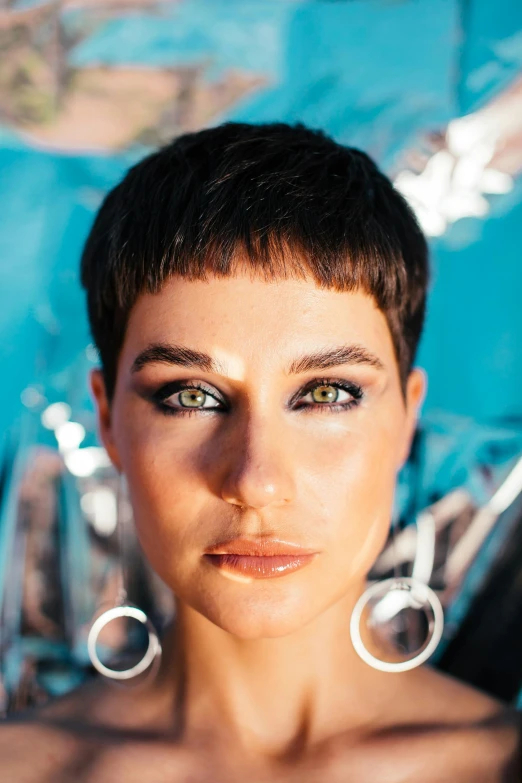 a woman posing for a picture in front of a blue background, an album cover, by Julia Pishtar, trending on pexels, short black pixie cut hair, high angle closeup portrait, clear green eyes, beachfront