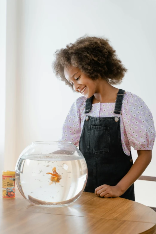 a little girl looking at a fish in a bowl, wearing a cute top, ashteroth, award - winning details, overalls