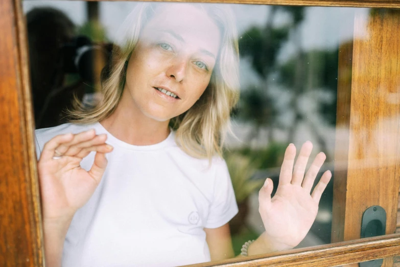 a close up of a person looking out a window, waving at the camera, a blond, dressed in a white t shirt, in a frame