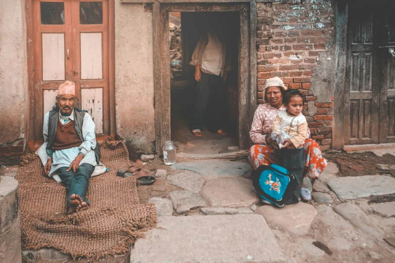 a group of people sitting in front of a building, pexels contest winner, in a village street, husband wife and son, thumbnail, josh grover