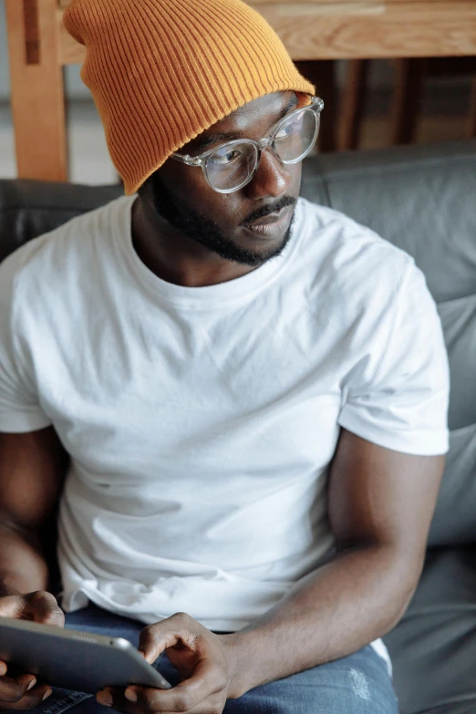 a man sitting on a couch using a tablet computer, an album cover, inspired by Terrell James, trending on pexels, wearing small round glasses, he also wears a grey beanie, man in white t - shirt, staring seductively