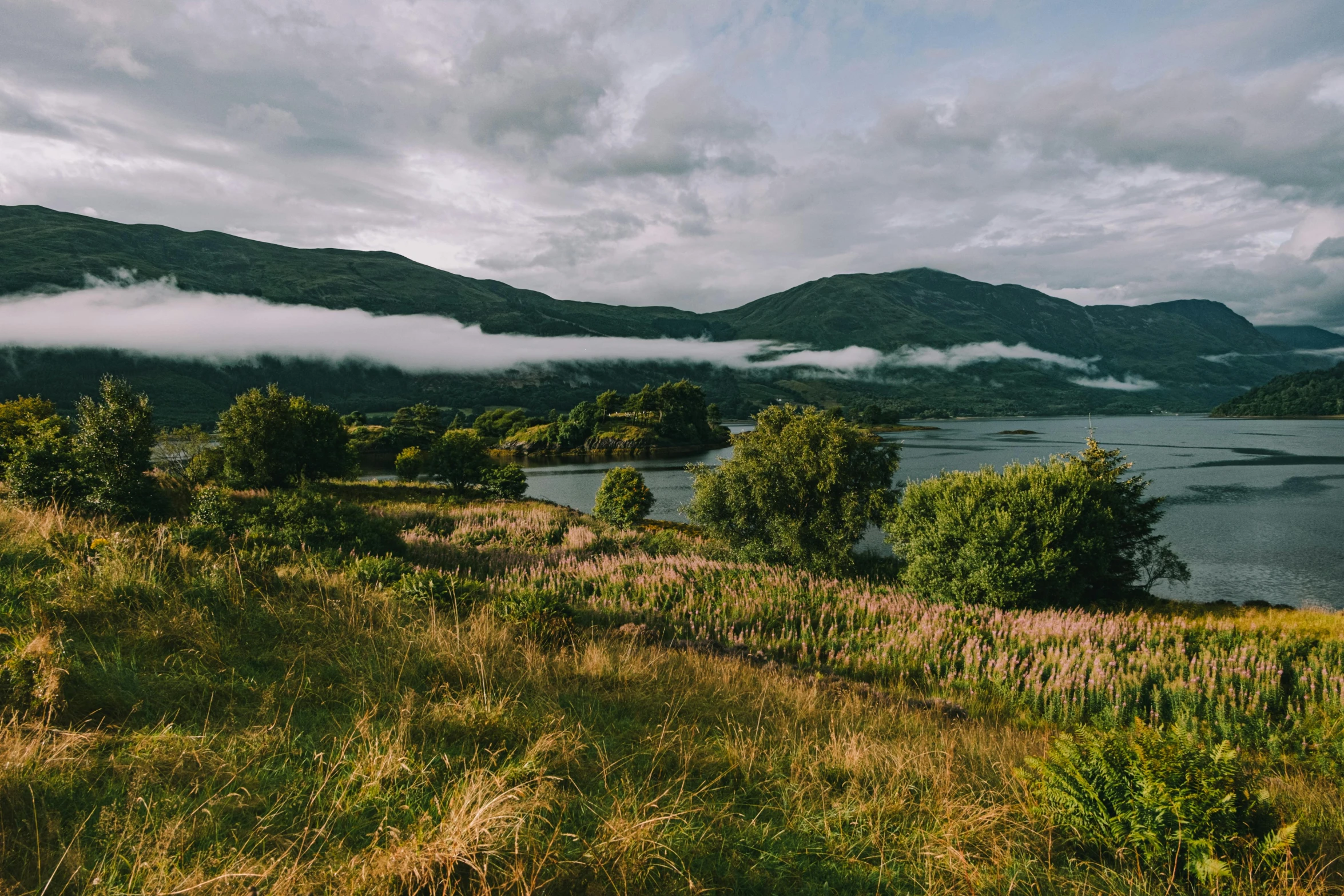 a large body of water sitting on top of a lush green hillside, by Jessie Algie, unsplash contest winner, hurufiyya, loch ness monster, loots of clouds, manuka, late summer evening