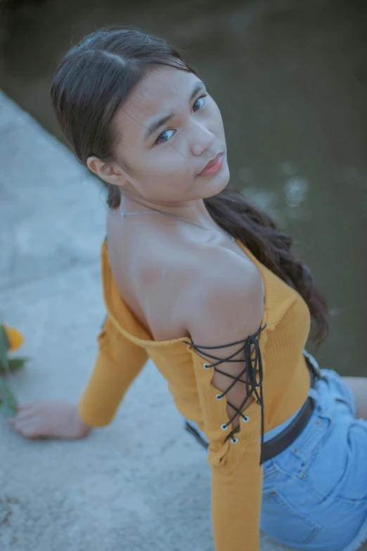 a woman sitting on a ledge next to a body of water, an album cover, by Reuben Tam, trending on pexels, renaissance, wearing yellow croptop, isabela moner, high angle close up shot, lovingly looking at camera