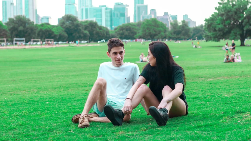 a couple of people sitting on top of a lush green field, in a city park, profile image, zachary corzine, teenager