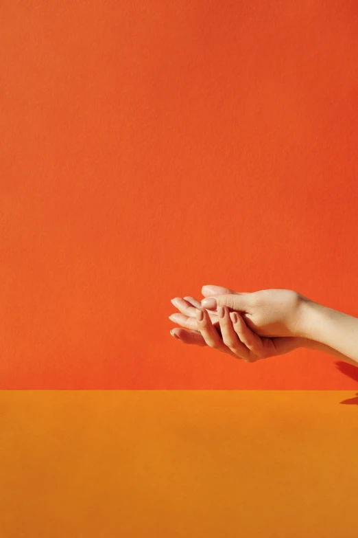 a woman holding her hands out in front of an orange wall, by Arabella Rankin, minimalism, david kassan, hand on table, promotional image, complementary colour