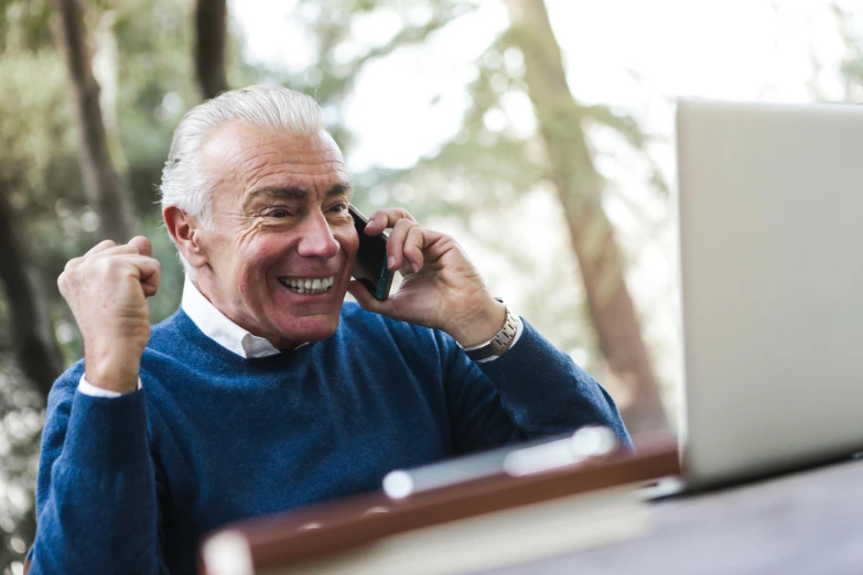 a man sitting in front of a laptop talking on a cell phone, a silver haired mad, laughing man, selling insurance, lush surroundings