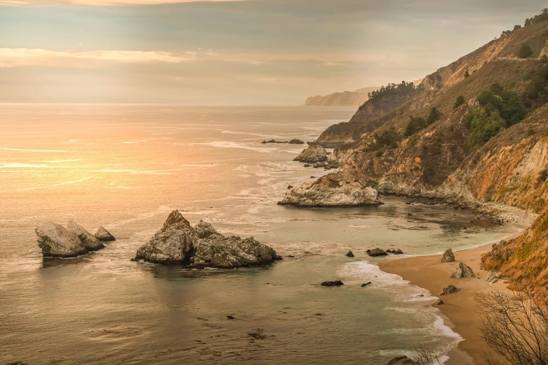 a group of rocks sitting on top of a beach next to the ocean, pexels contest winner, renaissance, golden hour in pismo california, steep cliffs, panoramic, hazy water