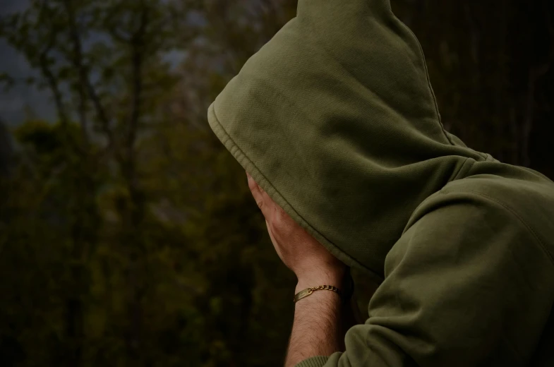 a man in a green hoodie covers his face with his hands, pexels contest winner, melancholy scenery, background image, trees in background, distant hooded figures