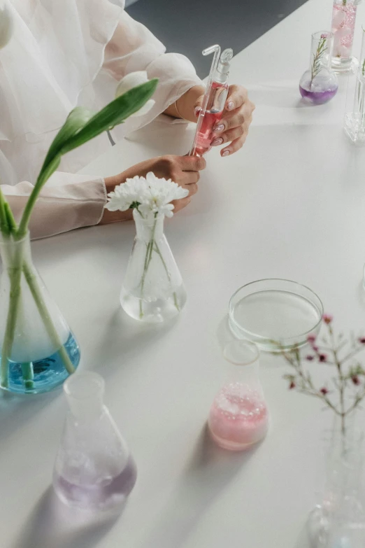 a woman sitting at a table with vases filled with flowers, trending on unsplash, process art, scientific glassware, wearing lab coat, pastel', detail shot