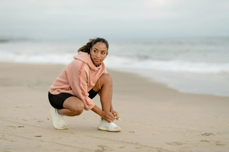 a woman squatting on the beach tying her shoe, pexels contest winner, happening, wearing a pastel pink hoodie, young black woman, chloe bennet, avatar image