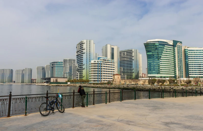 a bicycle leaning against a fence next to a body of water, high rise buildings, ameera al taweel, in a cybercity, portait image